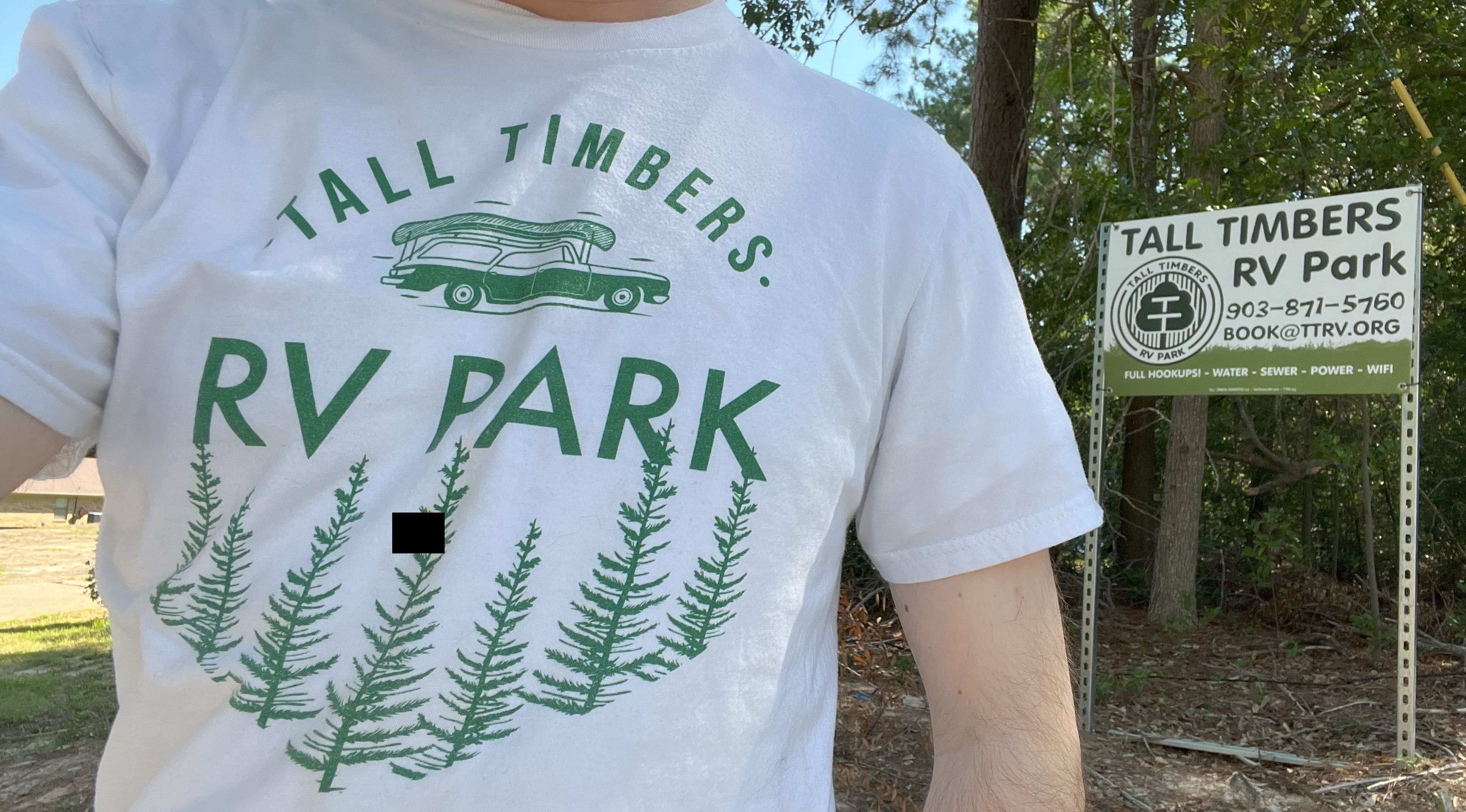 Customer wearing a Tall Timbers RV Park shirt standing by the main entrance sign, smiling on a sunny day